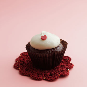 Chocolate cupcake with white frosting and a red heart on a red lace doily against a pink background