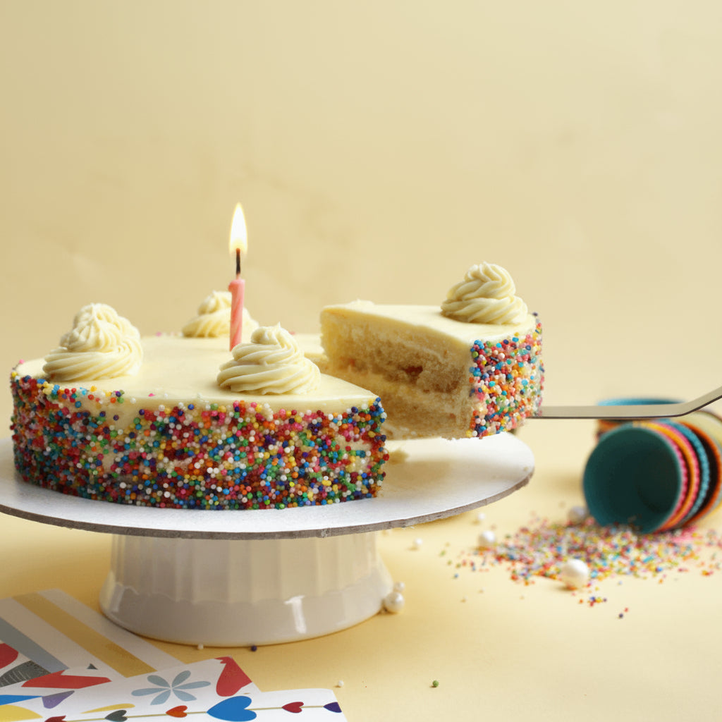 Birthday cake with a slice taken out, candle lit, on a white cake stand with colorful sprinkles.