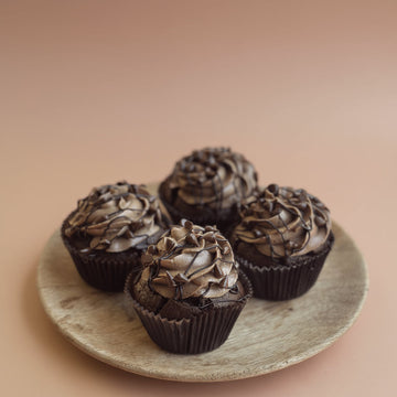 Four chocolate cupcakes with frosting on a wooden plate against a beige background