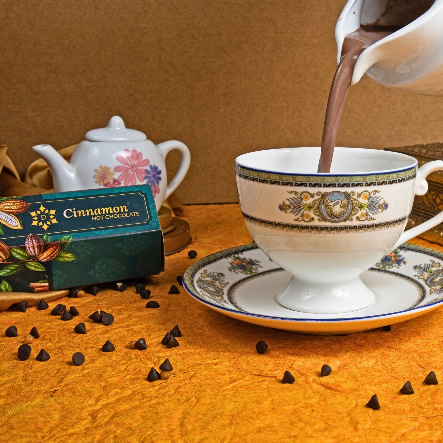 Cup of hot chocolate being poured with a box of cinnamon hot chocolate and chocolate chips on a wooden surface.