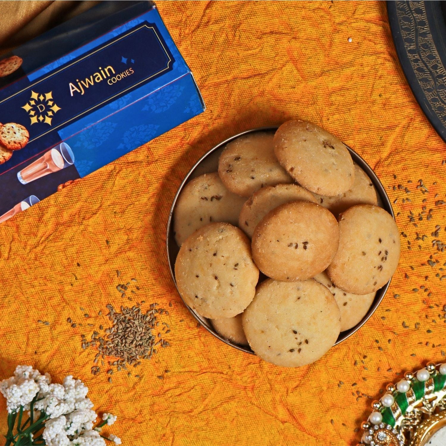 Ajwain cookies in a metal bowl on an orange fabric background with Ajwain cookies packaging.