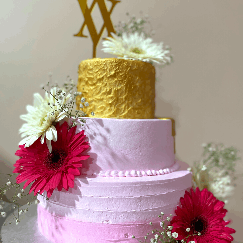 Three tiered wedding cake decorated with live flowers and its third layer wrapped in pretty edible gold foil and delectable stairs of whipped cream in shades of pink
