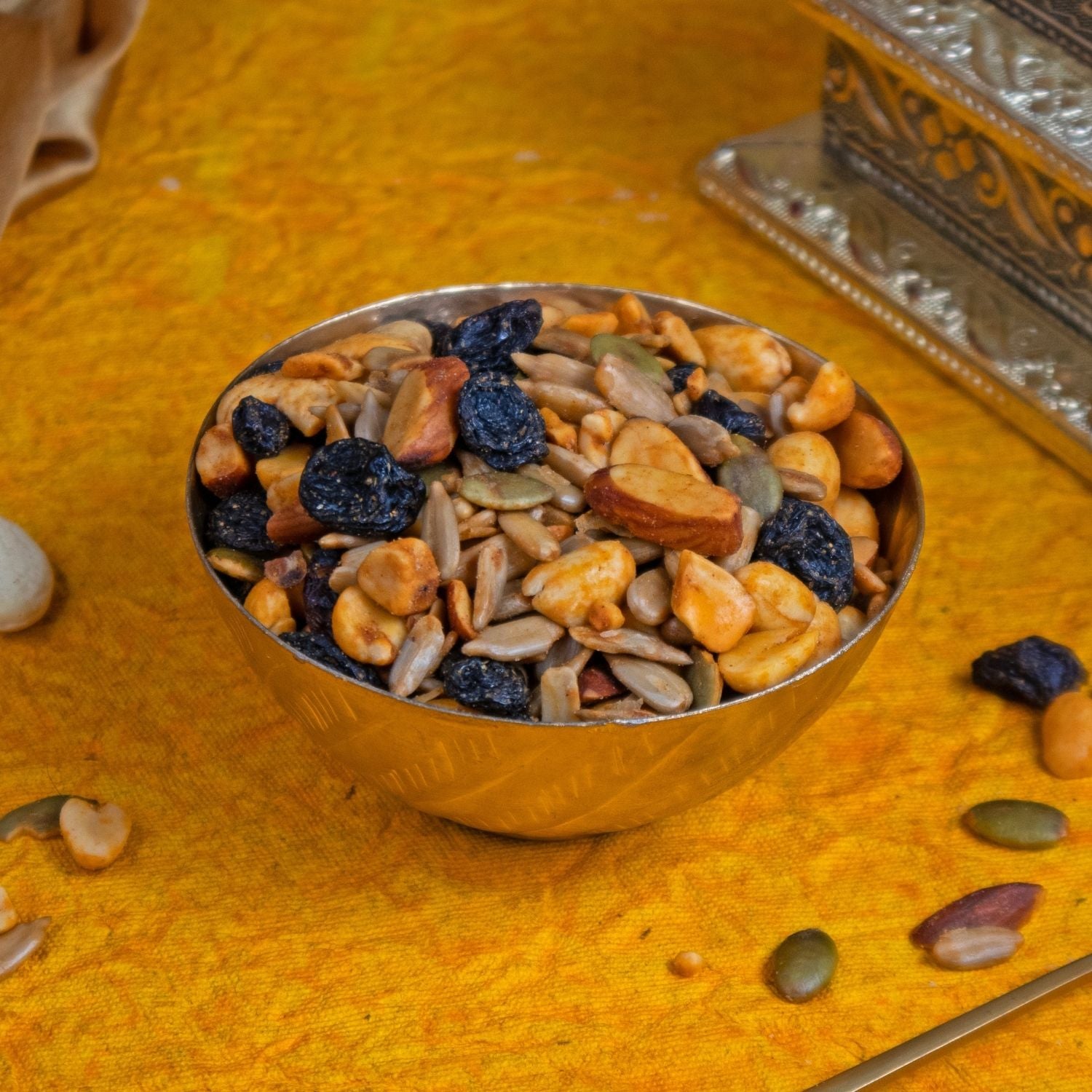 Metallic bowl filled with a mix of dried fruits and nuts on a textured yellow surface.