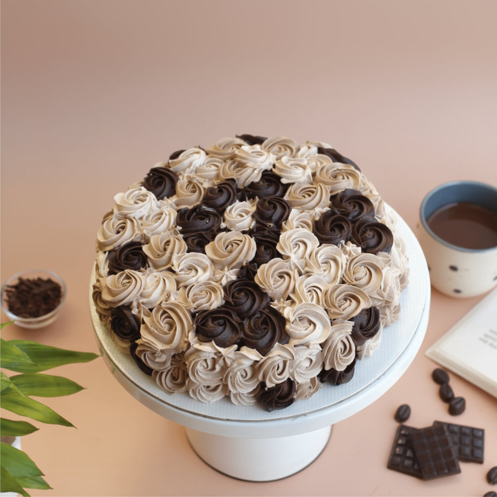 Decorative cake with black and white frosting on a stand, with a cup of coffee and chocolate pieces in the background.