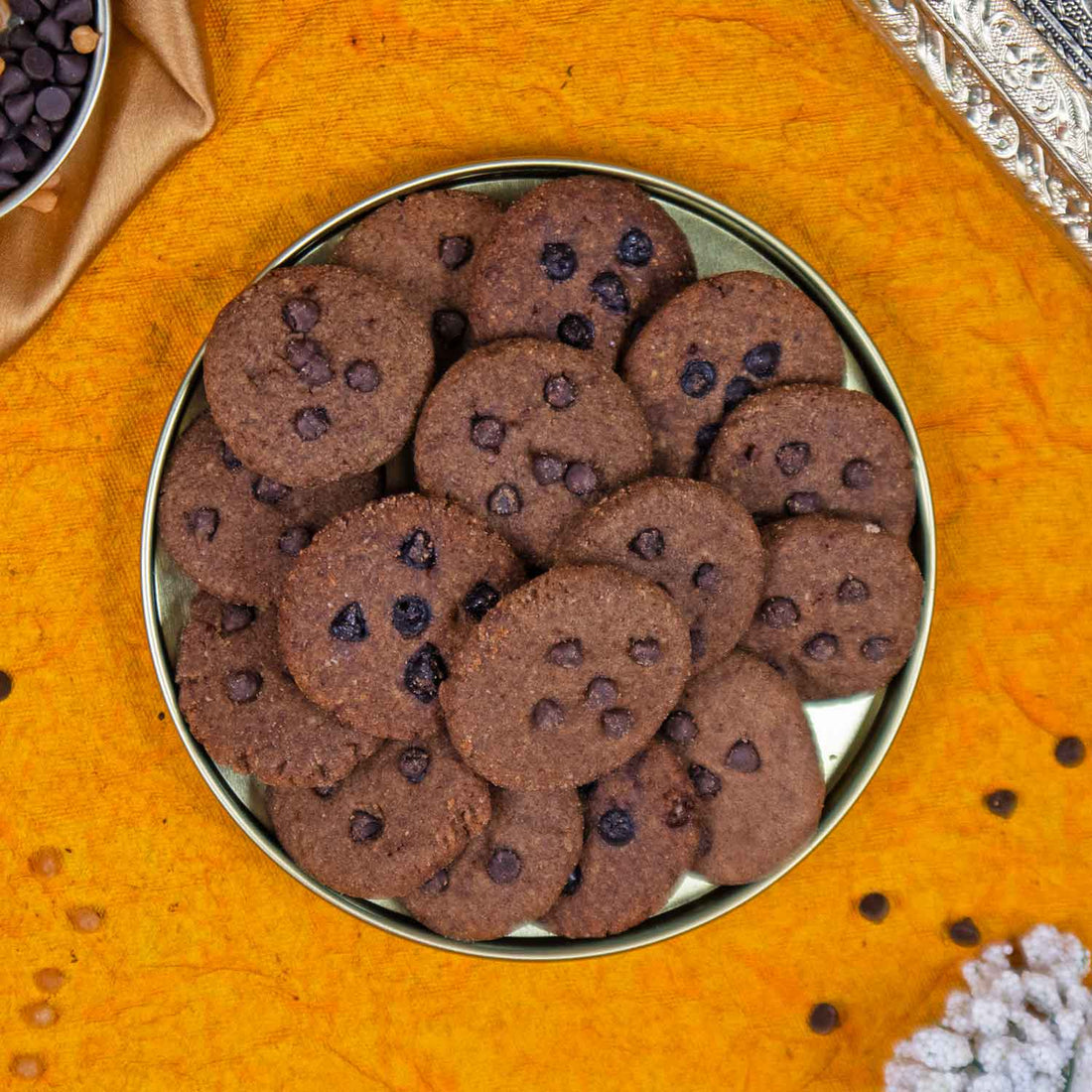 Ragi cookies with chocolate chips in a metal tin on an orange surface.
