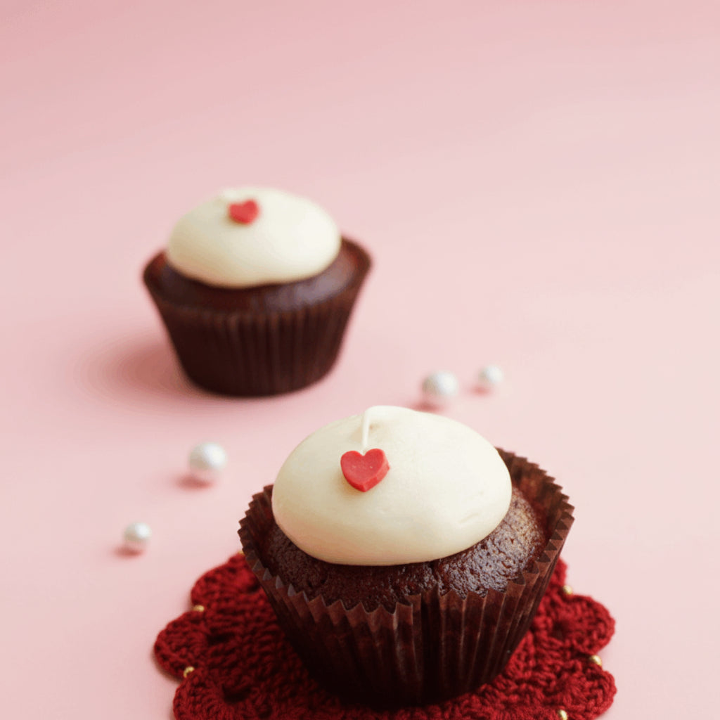 Chocolate cupcake with white frosting and a red heart on a red lace doily against a pink background