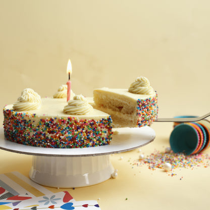Birthday cake with a slice taken out, candle lit, on a white cake stand with colorful sprinkles.