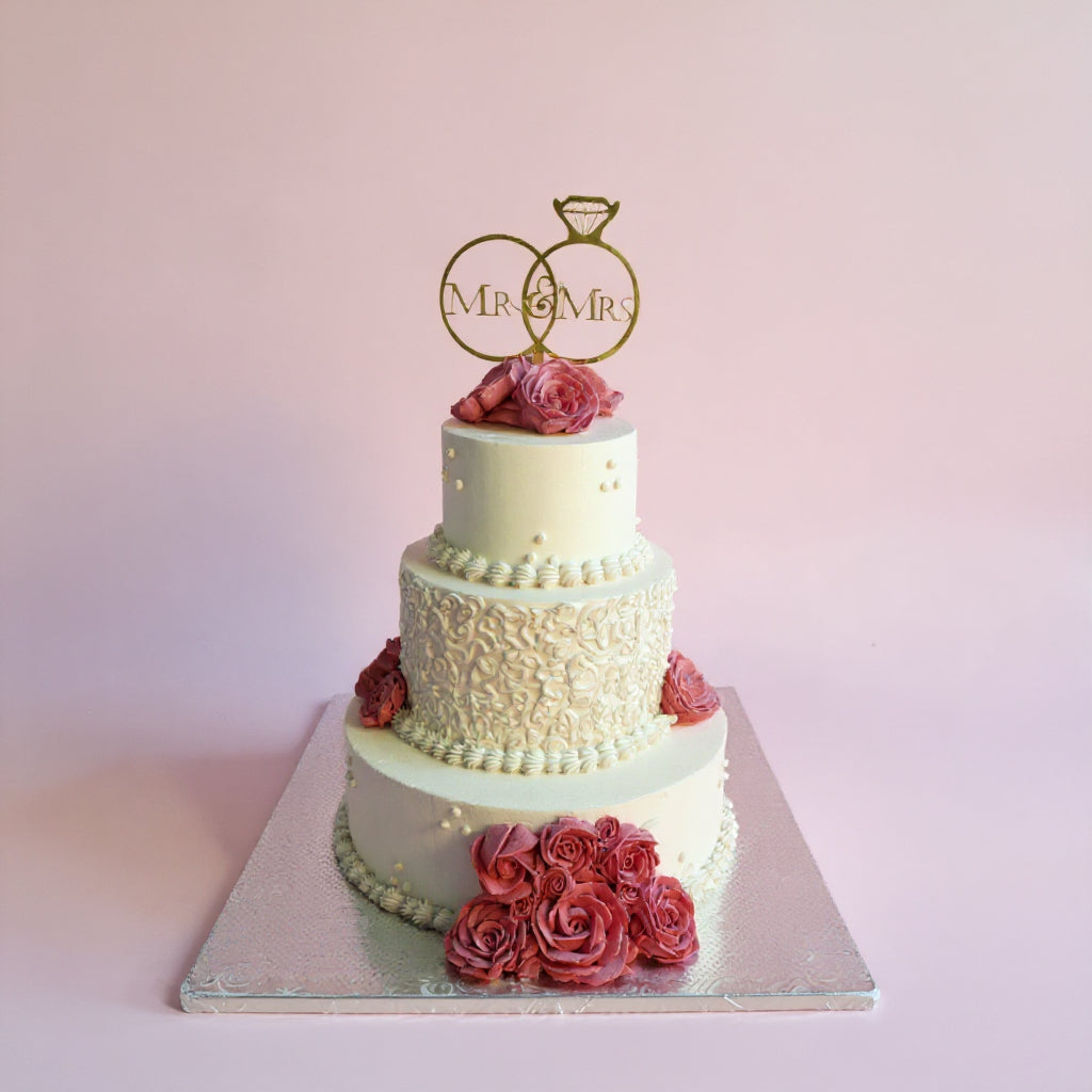 Three-tiered wedding cake with floral decorations and 'MR & MRS' topper on a light pink background