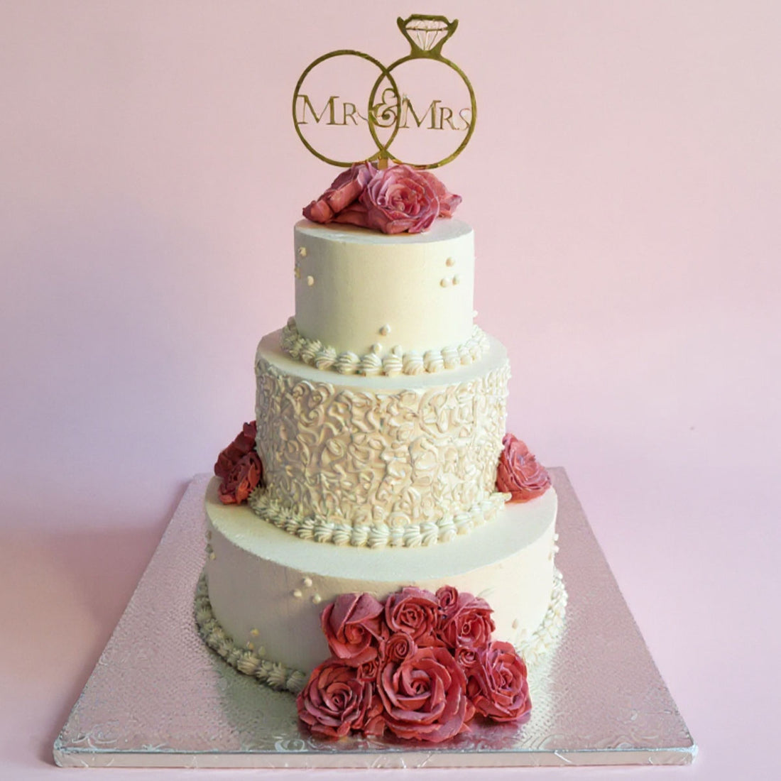 Three-tiered wedding cake with floral decorations and 'Mr & Mrs' topper on a light pink background