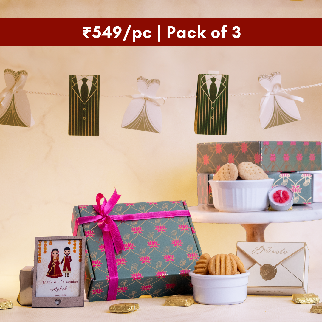 a wide shot of the golden moments hamper in the centre surrounded by a  customisable photoframe, a bowl of khara biscuits, a envelope box, a bowl of rava laddoo cookies and a floral tea light candle with chocolates wrapped in gold scattered around 
above the hamper bride and groom boxes hang on a thread
there are two more hampers stacked behind giving the picture a luxurious look 