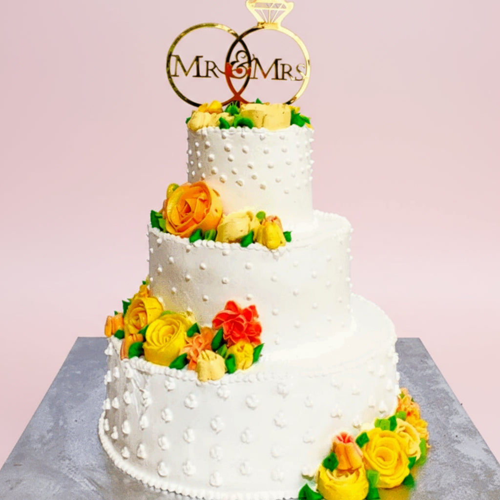Three-tiered wedding cake with floral decorations and 'MR & MRS' topper on a light pink background